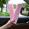 A hand holds two pink plush seat belt covers against a car window.