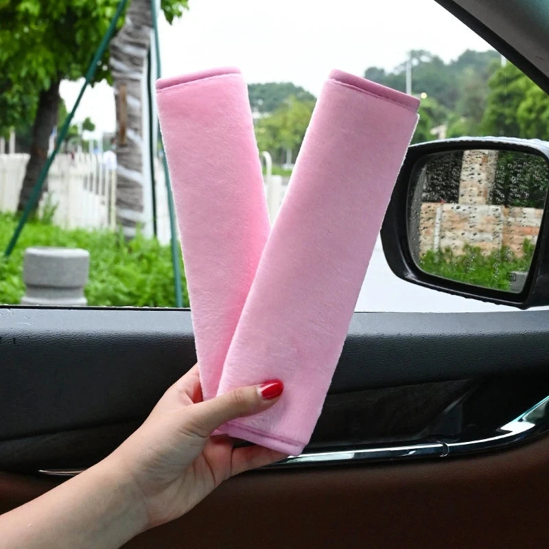 A hand holds two pink plush seat belt covers against a car window.
