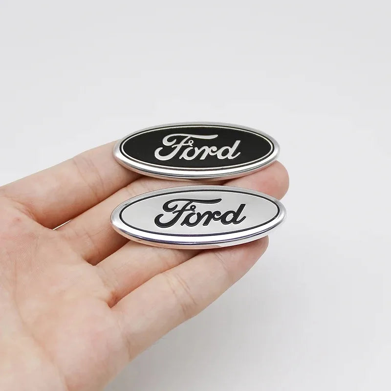 Eine Hand hält zwei ovale Ford-Lenkrad-Zieraufkleber, einer schwarz und einer silber.