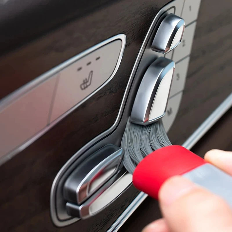 A hand is using a specialized brush to clean the air vent of a car interior.