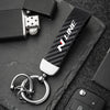 A close-up shot of a black carbon fiber style keychain with a silver split ring attached, resting on a dark slate surface.