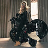A woman wearing black leather pants and sunglasses sits on a black motorcycle with a bell keychain.