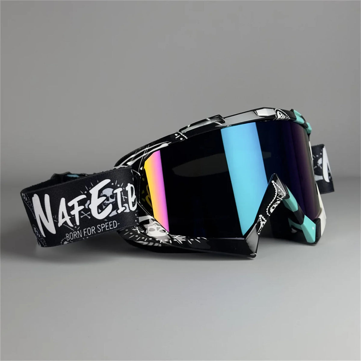 A pair of black motorcycle off-road goggles with a silver frame and colorful lenses resting on a gray surface.