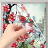 A hand touches a decorative PVC stained glass window film featuring pink and red floral designs on a tree branch.