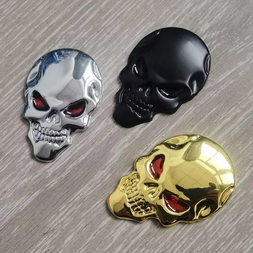 Three skull-shaped chrome emblems in silver, black, and gold on a gray surface.