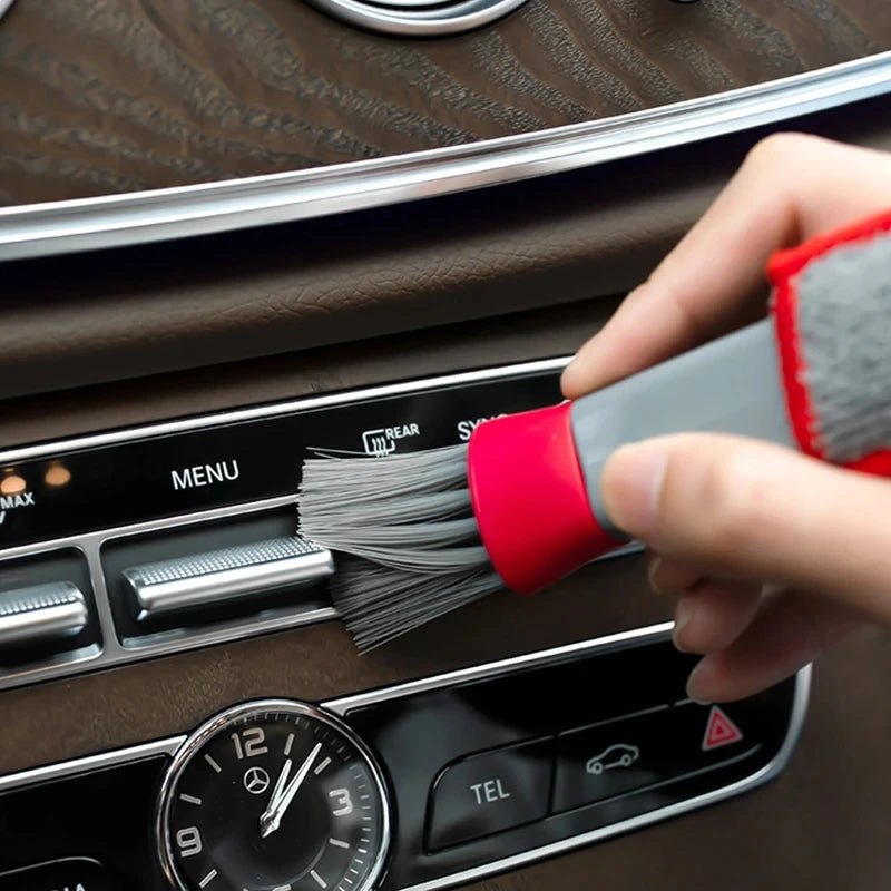 A hand uses a red and gray brush to clean the air vent of a car interior.