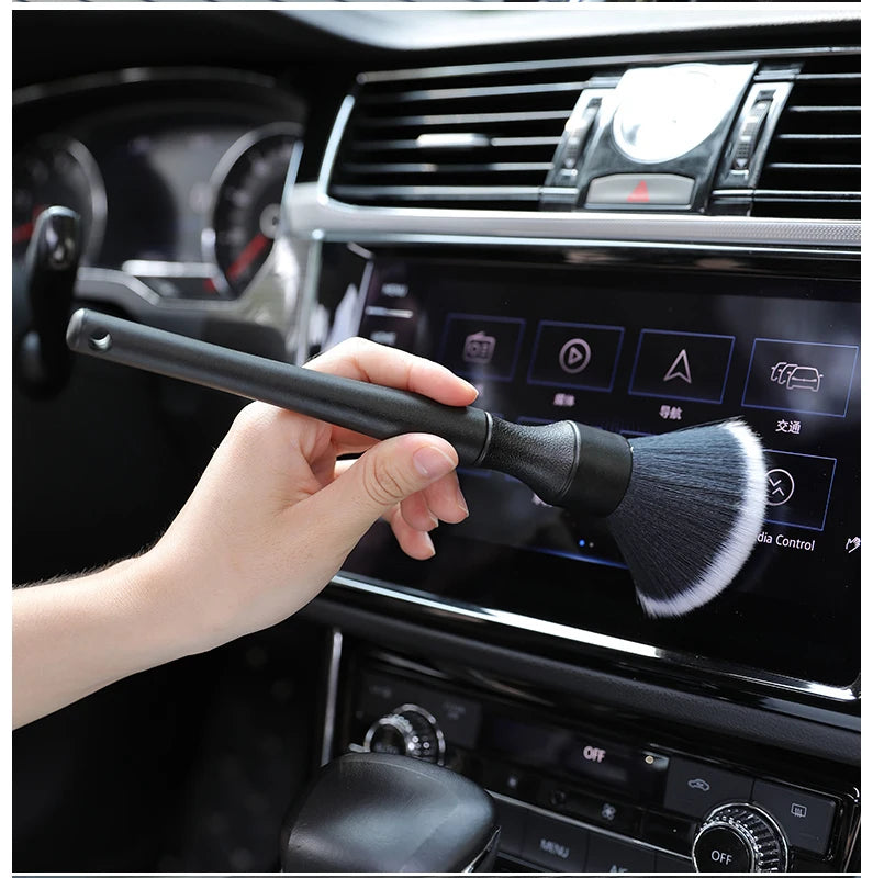 Une main tient un pinceau de détail pour intérieur de voiture, positionné près du tableau de bord et des bouches d'aération.