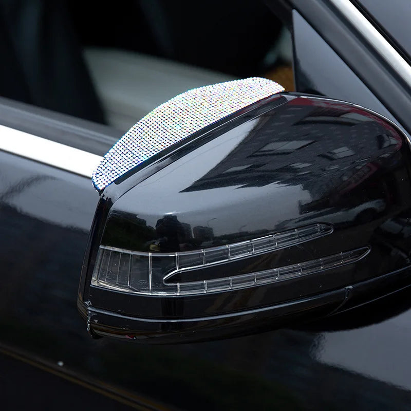 A close-up of a black car's rearview mirror with a clear plastic rain shield visor featuring sparkling rhinestones.