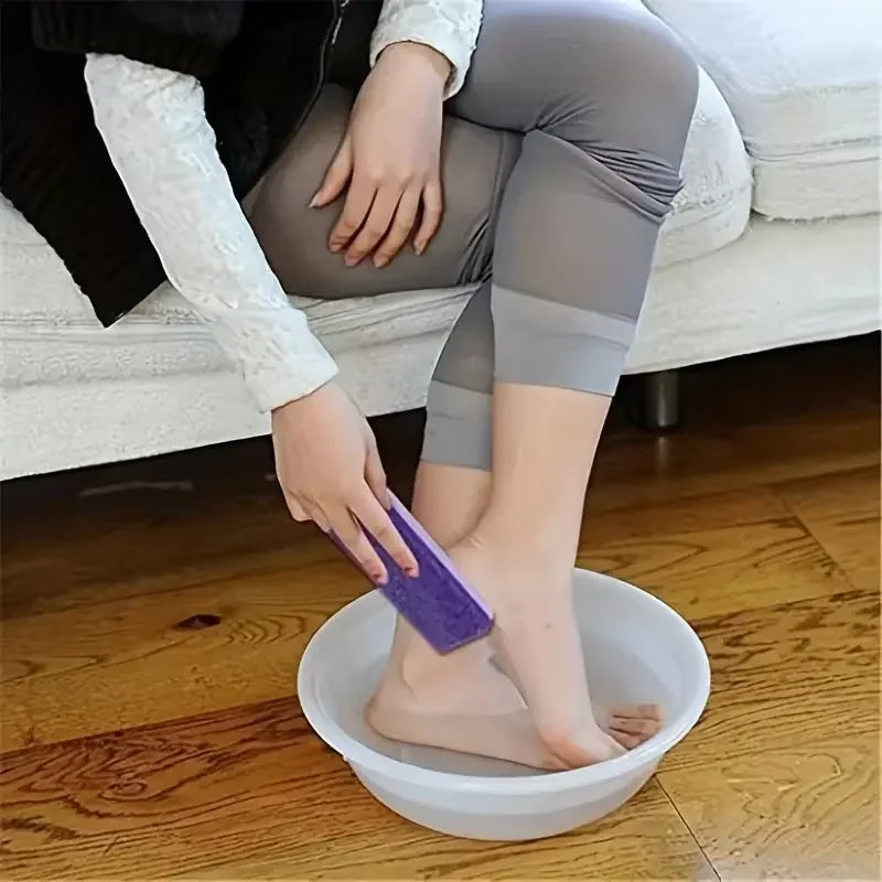 Une personne utilise une pierre ponçante en verre violet dans un bol blanc pour exfolier ses pieds.