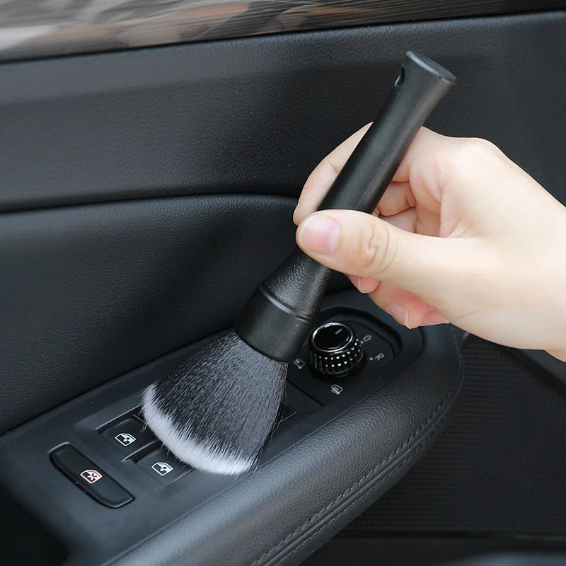 Une main tient une brosse de detailing noire à poils blancs, positionnée sur un outil de nettoyage d'intérieur de voiture.