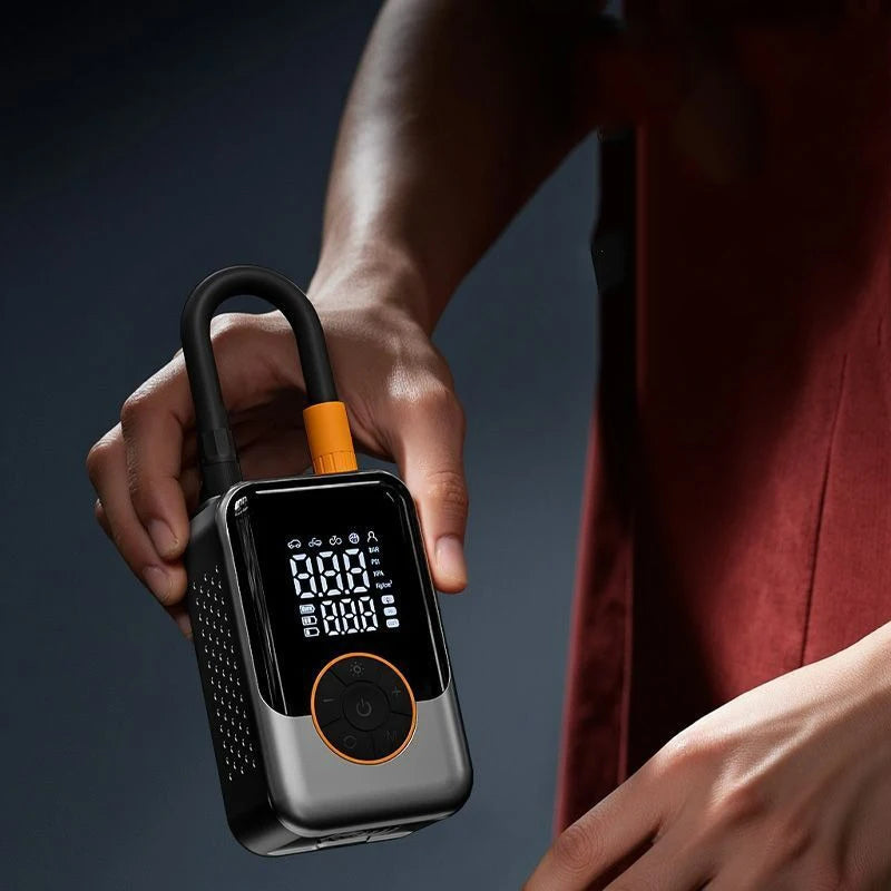 A hand holds a black wireless portable tire inflator with an orange button and digital display against a dark background.