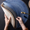 Hands are holding a stack of blue and beige woven cotton yarn placemats.