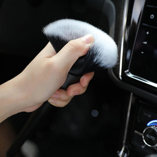 A hand holds a black detailing brush with soft white bristles against a dark car interior background.