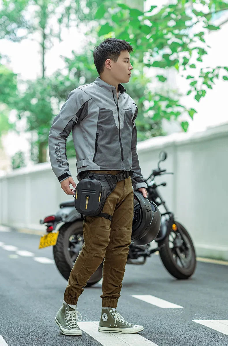 A young man wearing a gray jacket and brown pants stands next to a black motorcycle with a waterproof fanny pack.