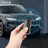 A hand holds a carbon fiber car key case cover near a modern dark gray car's front grille.