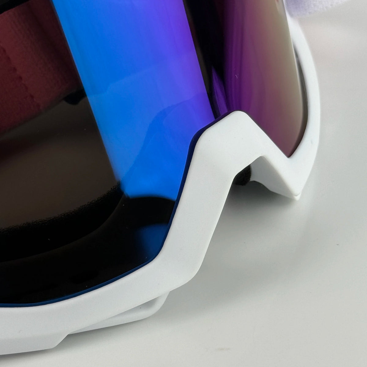 A close-up view of white off-road motorcycle goggles with blue and purple tinted lenses resting on a white surface.