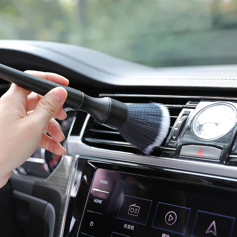 Une main tient une brosse de detailing noire près de la grille d'aération et des commandes d'une voiture intérieure.