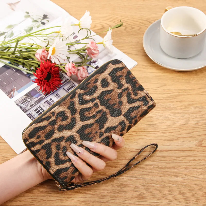 A leopard print long wallet is shown resting on a wooden table next to a cup and some flowers.