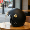 A black two-lens retro motorcycle helmet with a clear visor and gold accents resting on a wooden surface.