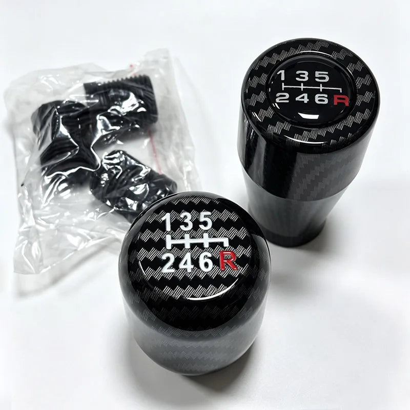 A black gear shift knob with the markings 135 246 R is shown next to a clear plastic bag containing spare knobs.