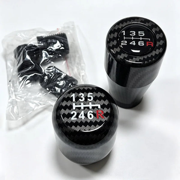 A black gear shift knob with the markings 135 246 R is shown next to a clear plastic bag containing spare knobs.