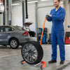 A man in blue overalls is holding an orange tire dolly with a large tire on a rolling cart in a garage setting.
