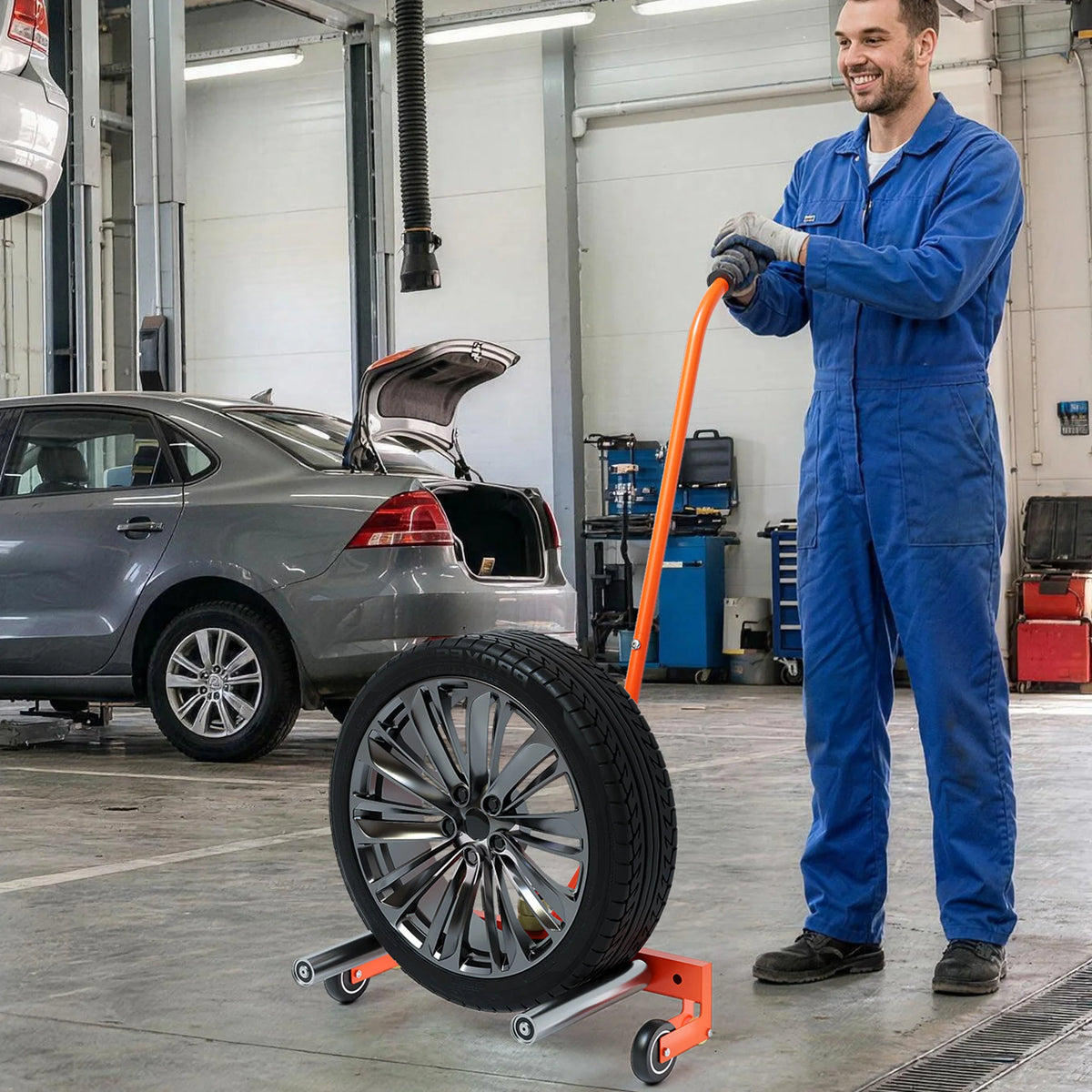 A man in blue overalls is holding an orange tire dolly with a large wheel in a garage setting.
