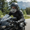 A man wearing a black leather jacket and helmet rides a motorcycle on a road with mountains in the background.