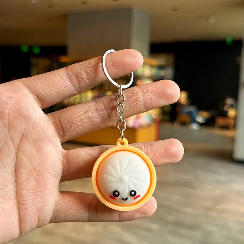 A hand holds a cute white and orange silicone keychain shaped like a bao bun with a smiling face.
