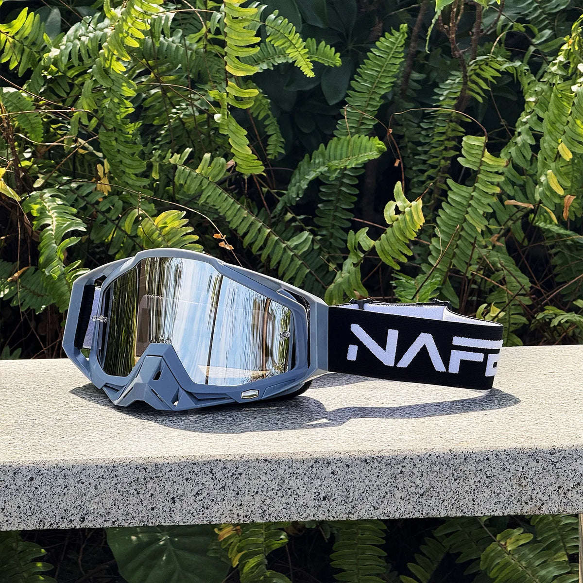 Une paire de lunettes de sport grises avec une sangle noire et le logo NAFF, posées sur une surface claire parmi le feuillage