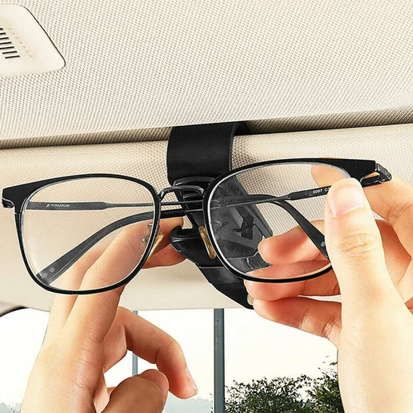A hand is holding a black clip designed to hold eyeglasses or sunglasses, positioned near a car window.