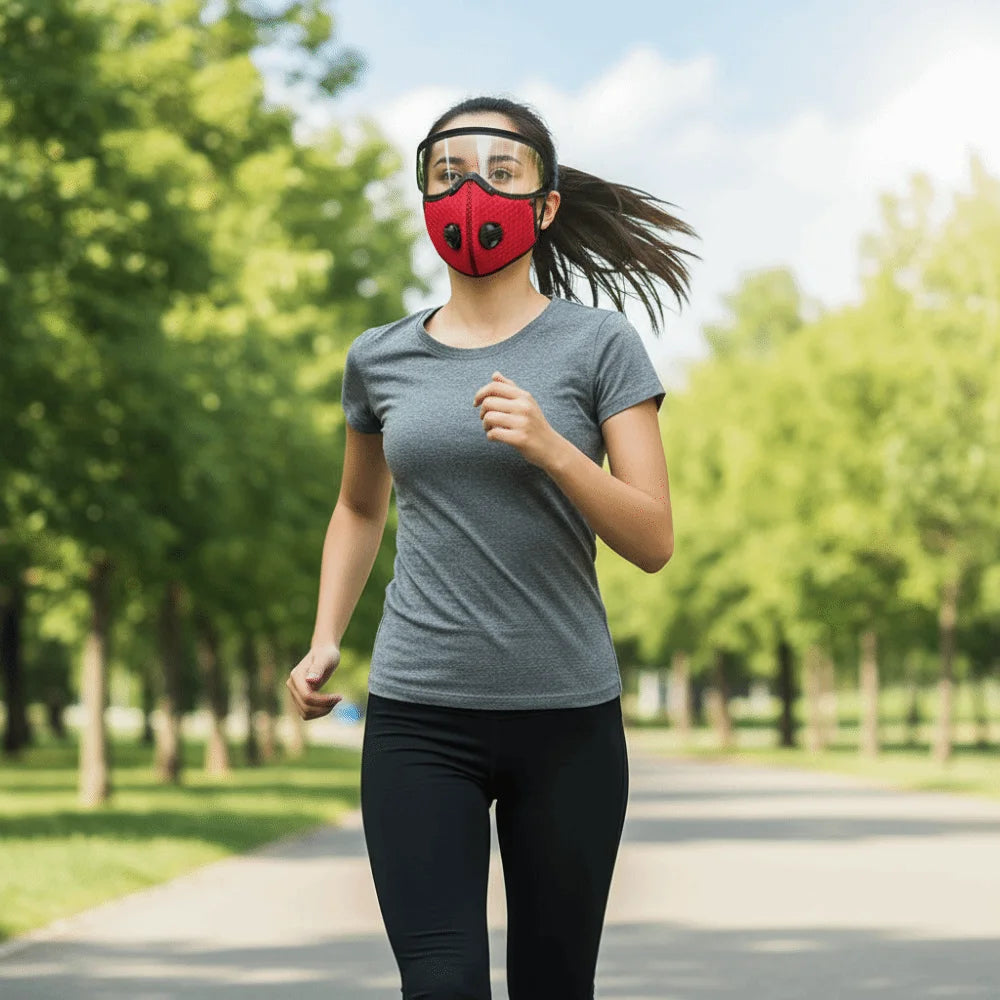 A woman wearing a red face mask and protective goggles is jogging outdoors on a paved path surrounded by green trees.