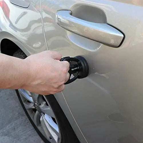 A hand is using a car dent remover puller with a suction cup to lift a panel on a vehicle.