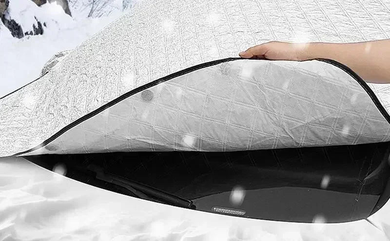 A hand is holding a black universal car windshield sunshade and snow protector against a white surface in snowy conditions.