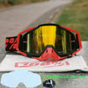 A pair of orange and black motocross goggles resting on a white cloth, with a graphic showing 100% clear lens.