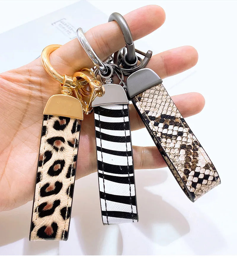 Una mano sostiene tres llaveros de cuero con estampado de serpiente leopardo y herrajes de oro y plata.