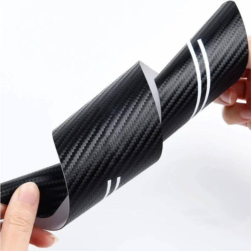 A close-up of a black carbon fiber door sill protector with white stripes being held by a hand.
