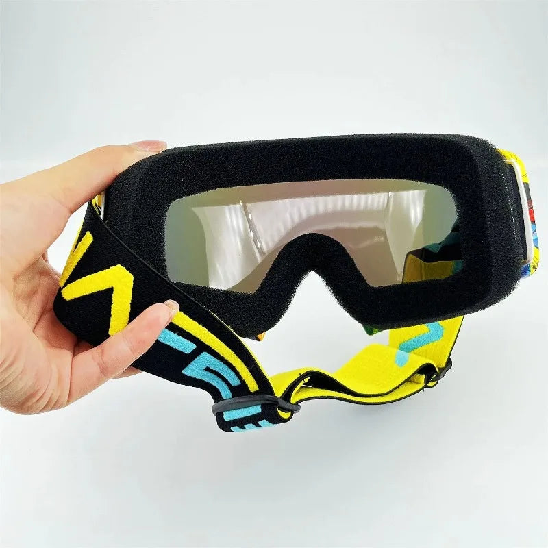 Una mano sostiene unas gafas de motocicleta y moto de tierra todo en uno, negras y amarillas, con lente transparente.