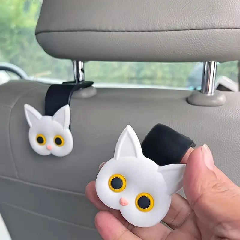 A hand holds a white 3D cartoon cat headrest hook designed for universal storage.