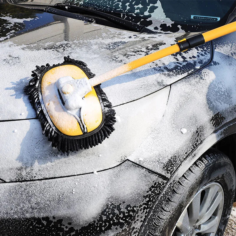 A yellow telescoping car wash mop with a black handle is cleaning snow off a dark vehicle.