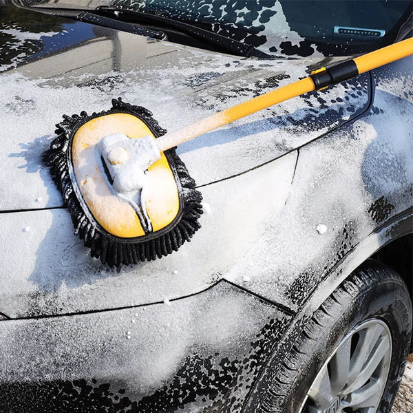 A yellow telescoping car wash mop with a black handle is cleaning snow off a dark vehicle.