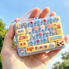 A hand holds a clear plastic case filled with colorful cartoon keycap stress relief buttons and a pink string.