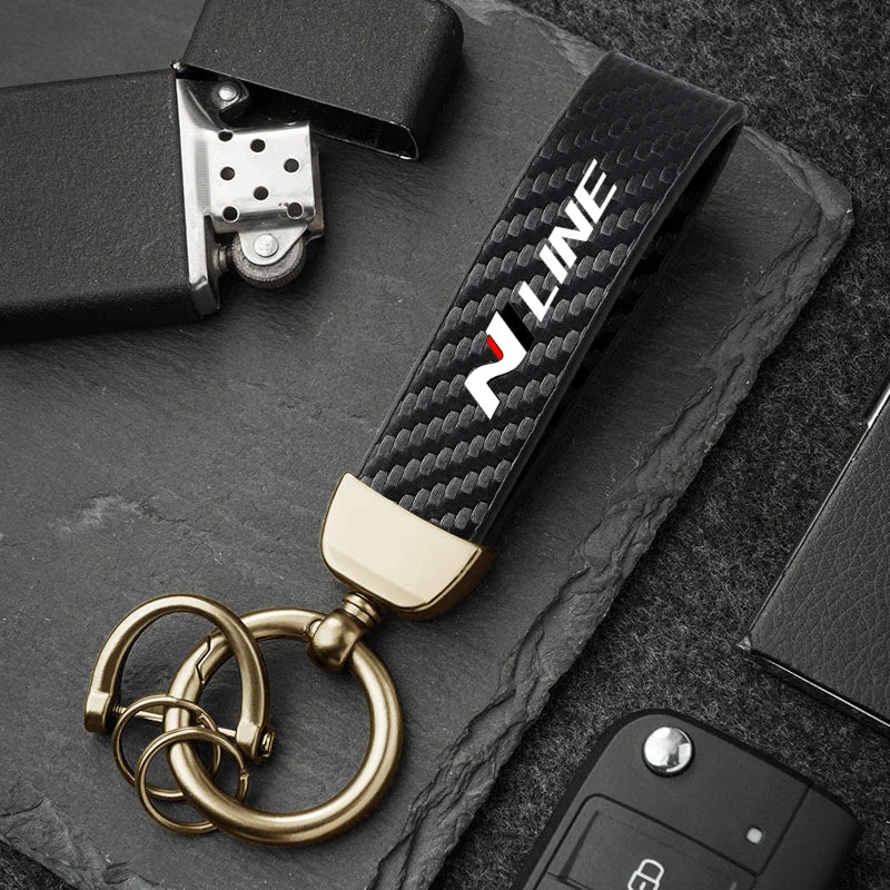 A close-up of a black and gold luxury carbon fiber style keychain with a silver clasp resting on a dark slate surface.