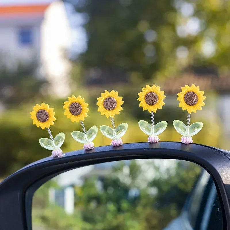 Ornements de tournesols jaunes avec tiges vertes et accents roses, présentés sur une console ou un miroir de voiture.