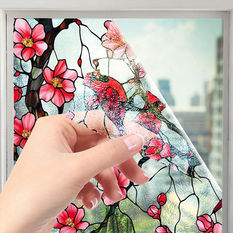 A hand touches a decorative PVC stained glass window film featuring pink and red floral designs on a tree branch.
