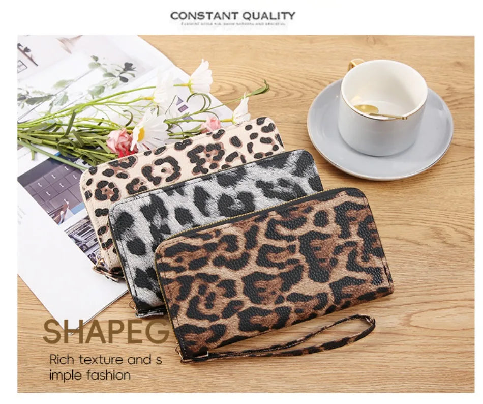A close-up of a brown leopard print long wallet resting on a wooden surface next to a white cup and some greenery.