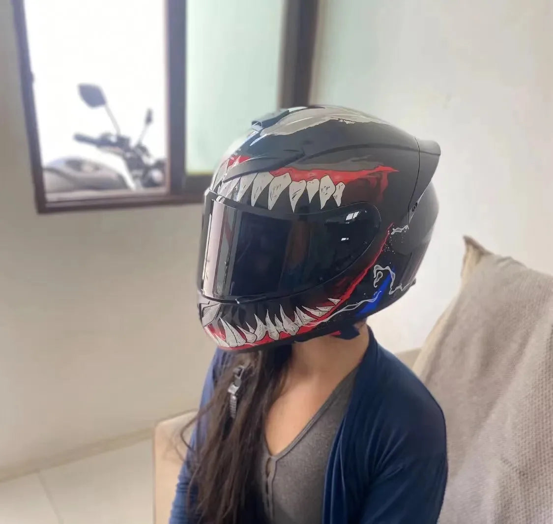 A person is wearing a black full face motorcycle helmet with a fierce, toothy design against a light background.