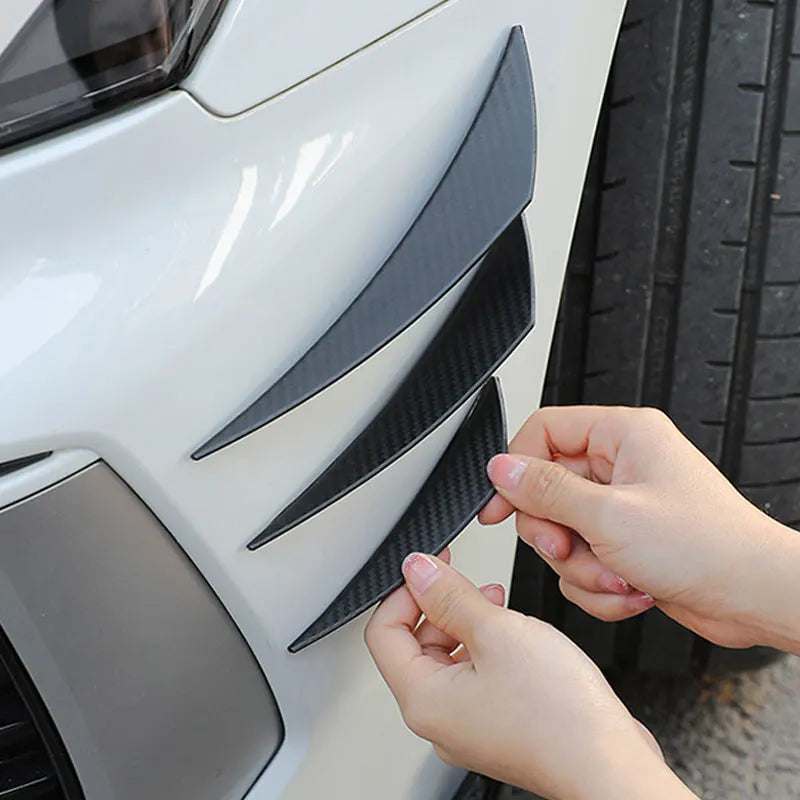 A hand is adjusting black universal car spoiler fins on a white vehicle's front bumper.