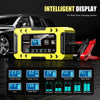 A yellow portable 12V smart battery charger device with a display and red clamp is shown on a dark surface.
