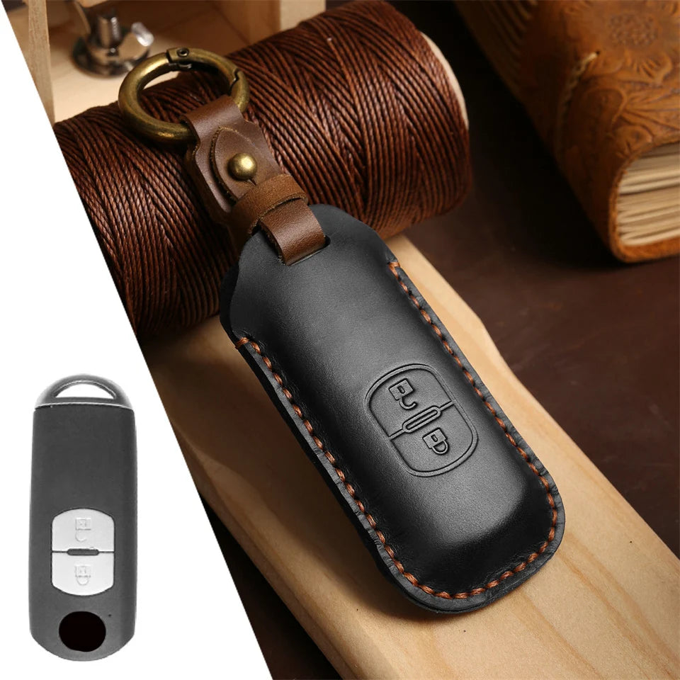 A black leather car key fob cover attached to a brass ring, resting on a wooden surface next to a rolled-up brown leather str